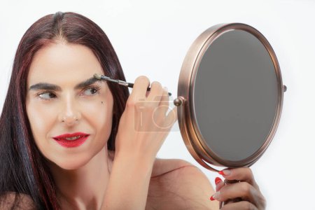 Photo for Adult woman applying makeup while looking at herself in the mirror against a white background - Royalty Free Image