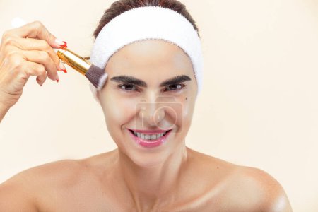 Photo for Smiling woman's face applying makeup with a brush in hand against a light background - Royalty Free Image
