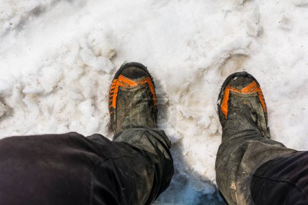 Téléchargez les photos : Vue de dessus des bottes de randonnée sur les pieds d'un pantalon touristique et souillé lors d'une randonnée en montagne dans la neige de printemps. - en image libre de droit