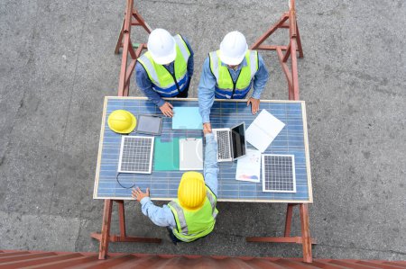 Blick von oben auf professionelle Ingenieure, die Helme und Warnwesten tragen und sich mit Solarmodulen zum Händedruck treffen, um das Projekt zu vereinbaren. Konzept für grüne alternative Energien