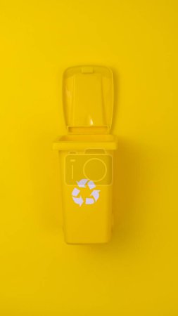 Papelera de reciclaje amarilla con tapa abierta sobre fondo amarillo a juego, que simboliza la gestión de residuos y el cuidado del medio ambiente.