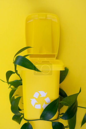 A yellow trash can sits adjacent to a vibrant green plant, creating an interesting contrast between man-made waste and the natural environment.