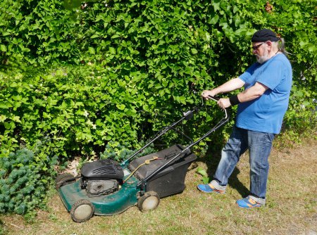 Téléchargez les photos : Homme âgé actif tondre l'herbe au soleil avec une tondeuse à gaz - en image libre de droit
