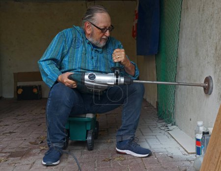 Foto de Hombre de edad avanzada activa está perforando un agujero en la pared de un garaje con un martillo neumático y un mordisco de núcleo de martillo giratorio - Imagen libre de derechos