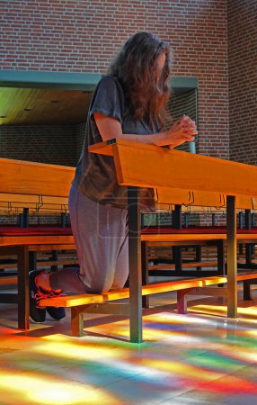 Foto de Una mujer arrodillada en oración en una iglesia. La luz del sol fluye a través de las vidrieras y pinta un patrón colorido en el suelo - Imagen libre de derechos