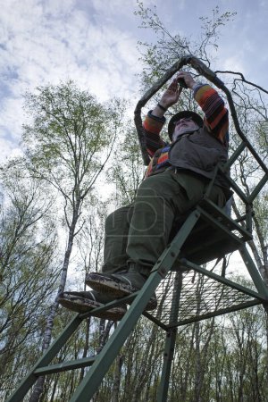 Téléchargez les photos : Un homme costaud s'assoit dans le haut stand d'un chasseur dans une belle tourbière et prend des photos - en image libre de droit