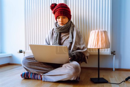 Teenagermädchen neben Heizkörper geht mit Laptop ins Internet, sitzt in warmem Schal, Mütze und gestrickten Wollsocken auf Parkettboden. erhitzt in der Nähe von Wärmequelle. Kühlhaus-Konzept, Wintersaison