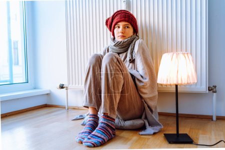 teenage girl in warm clothes, scarf and hat, sits near heating radiator on parquet floor at home, legs crossed, she warms up in cold house