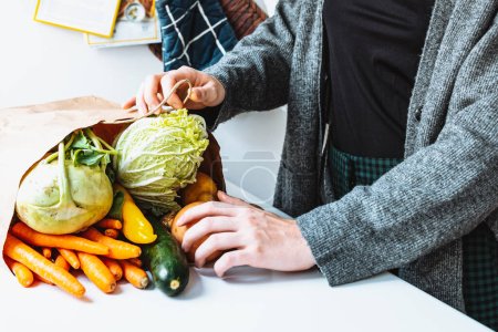 Photo for Fresh vegetables in eco bag on kitchen table. woman's hands take out fresh cabbage from paper bag. Delivery of products home. Illegal second-rate products for free - Royalty Free Image