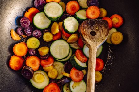 Foto de Verduras frescas multicolores rebanadas en círculos en una sartén vieja con cuchara de madera. Cena vegetariana cacerola de verduras, zanahorias de colores, calabacín, vista superior - Imagen libre de derechos