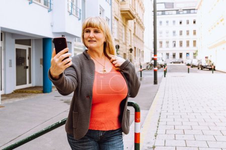 Photo for Middle-aged blonde busty woman takes selfie on smartphone camera, standing near house, against blurred background of residential modern area. Portrait of attractive middle age woman taking selfie in city - Royalty Free Image