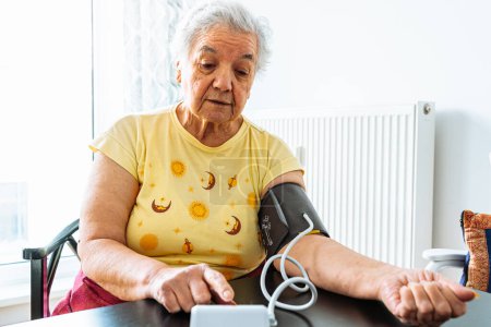 Téléchargez les photos : Elderly woman with hypertension measures her blood pressure at home. Sad overweight mature lady measures blood pressure with tonometer. Daily health check. Daily health monitoring, elderly life at home - en image libre de droit