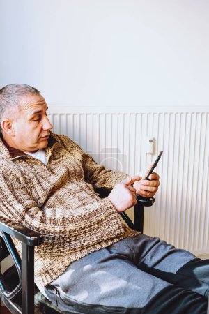 Téléchargez les photos : Middle-aged man with big fat belly, in warm knitted sweater, sits on chair near heating radiator, rummages through mobile Internet, concept of early aging and depression in middle age men - en image libre de droit