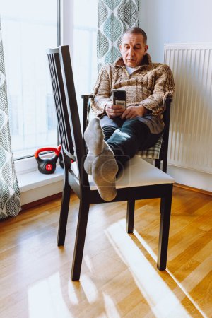 Photo for Middle-aged man with large fat belly sits on chair near heating radiator, with feet on another chair, against background of window and nearby sports weights. concept of early aging and depression in middle age men - Royalty Free Image