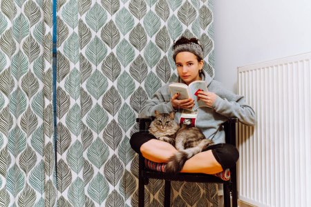 Téléchargez les photos : Teenager girl in home casual clothes sits on chair with legs crossed, with beloved cat together, reads book. Spending free time with your beloved pet home - en image libre de droit
