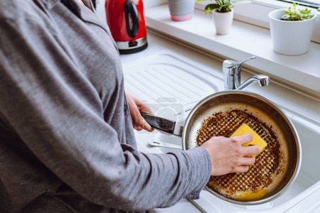 Photo for Woman's hand washes burnt greasy frying pan with kitchen washcloth in sink. Dirty dishes with burnt food, household chores, washing dishes - Royalty Free Image