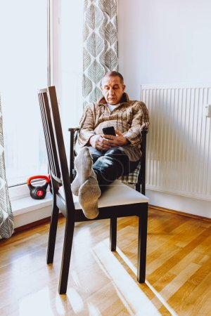 Photo for Middle-aged man with large fat belly sits on chair near heating radiator, with feet on another chair, against background of window and nearby sports weights. concept of early aging and depression - Royalty Free Image