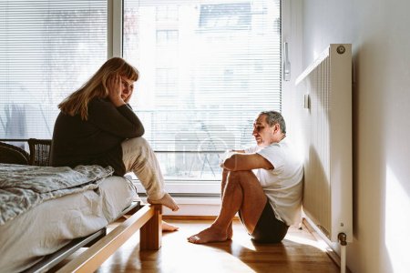 Foto de Middle-aged married couple is sitting in bedroom after quarrel. man sits on floor with legs crossed, woman sits on bed with hands behind head. Midlife crisis, family breakdown, quarrel or divorce - Imagen libre de derechos