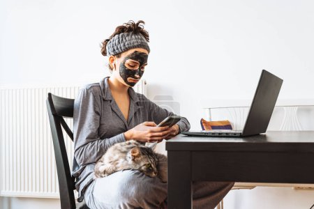 young girl in pajamas, cosmetic mask, with bandage on hair, sits at table in morning, in front of computer, domestic cat sits on lap. Home life with pets