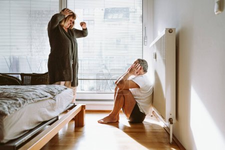 Family quarrel, showdown between spouses, scene of jealousy in bedroom. middle-aged married couple, man in underwear sits on floor and holds head, woman is scandalous, jealous, emotionally