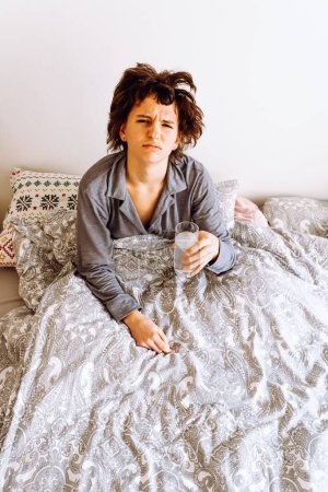 Téléchargez les photos : Teenage girl sits in bed, with shaggy hair from fatigue, sad face, holds glass of water and pill in hand - en image libre de droit