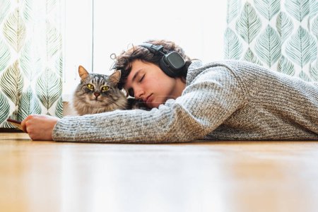 Photo for Spend your free time at home with your cat. teenage girl in headphones, with mobile phone, lies on floor in living room near large window, with fluffy Maine Coon cat, listens to music, closing eyes - Royalty Free Image