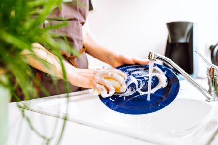 mujer en delantal de chefs marrón de trabajo, lava platos con detergente con esponja, en fregadero en cocina. concepto de limpieza, lavar platos, lavavajillas de trabajo