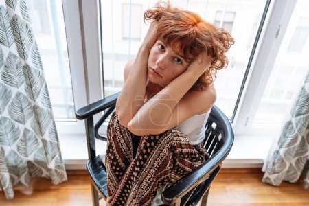 Portrait attractive red-haired teenage girl, in home clothes, sitting on chair in living room near large window.