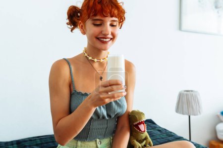 attraktives Teenager-Mädchen mit roten Haaren sitzt auf dem Sofa im Zimmer und trinkt frische, laktosefreie Milch auf pflanzlicher Basis aus Glas.