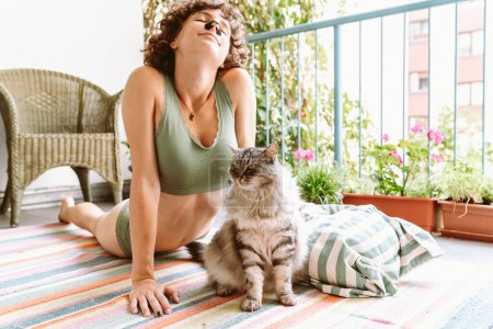 Téléchargez les photos : Curly jeune fille attrayante fait du yoga, relaxant, assis sur le balcon en haut vert, avec chat bien-aimé. Loisirs avec concept chat - en image libre de droit