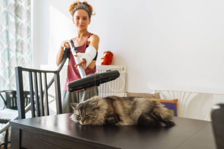 Téléchargez les photos : Adolescente aspirant chat couché sur la table dans la salle à manger. concept de lutte contre les poils d'animaux, nettoyage appartement - en image libre de droit