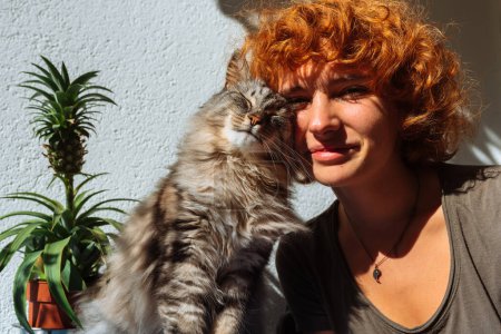 Téléchargez les photos : Portrait jeune fille aux cheveux roux attrayant et chat ronronnant moelleux dans les rayons du soleil, avec des ombres dures, étreignant tout en étant assis sur le balcon - en image libre de droit