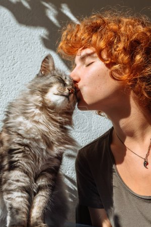 Téléchargez les photos : Portrait jeune fille aux cheveux roux attrayant et chat ronronnant moelleux dans les rayons du soleil, avec des ombres dures, étreignant tout en étant assis sur le balcon - en image libre de droit
