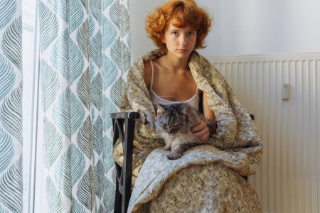 Mädchen umarmt Katze, sitzt in Decke zu Hause, gefroren. jugendlich barfuß, rothaarig, lockig, morgens, in Decke gewickelt, sitzt umarmende Hauskatze, neben Heizkörper, großes Fenster mit Vorhängen bedeckt.
