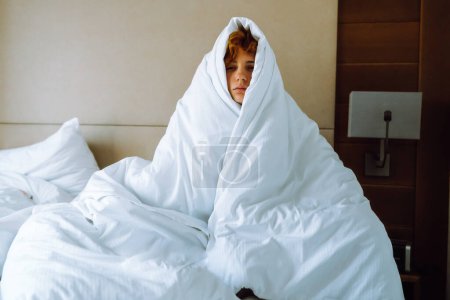 portrait adolescent assis sur le lit enveloppé dans une couverture, réveillé tôt le matin, déprimé, fatigué