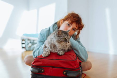 Porträt eines rothaarigen Teenagers, der mit einem Koffer und einer grauen, flauschigen Katze in einer leeren Wohnung auf dem Parkettboden sitzt. Konzept Umzug, Miete, Studentenleben