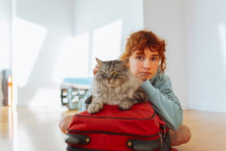 Téléchargez les photos : Portrait adolescente rousse assise sur le parquet, avec valise, avec chat moelleux gris, dans un appartement vide. concept déménagement, loyer, vie étudiante - en image libre de droit