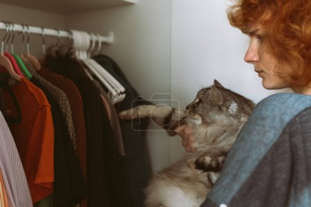 Red-haired curly teenage girl holds large gray Maine Coon cat in arms, choosing things from closet. spending time with pet