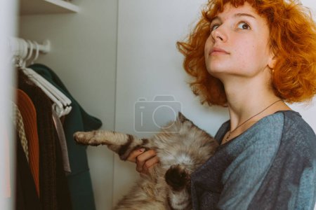 Red-haired curly teenage girl holds large gray Maine Coon cat in arms, choosing things from closet. spending time with pet