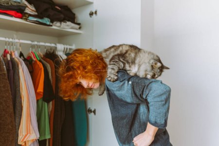 Red-haired curly teenage girl holds large gray Maine Coon cat in arms, choosing things from closet. spending time with pet. cat sits funny on girls shoulders