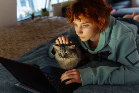 adolescente, en sweat-shirt, rousse, passe du temps avec le chat, couchée sur le lit, en utilisant un ordinateur portable, le soir