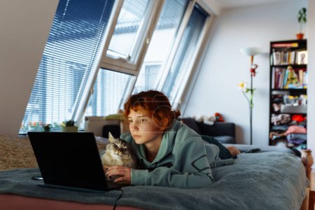 adolescente, en sweat-shirt, rousse, passe du temps avec le chat, couchée sur le lit, en utilisant un ordinateur portable, le soir
