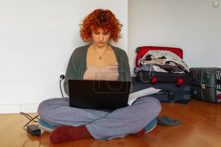 Porträt rothaarige Teenagerin, die auf dem Boden in einem leeren, unmöblierten Zimmer sitzt, Tablet benutzt, unmontierte Koffer in der Nähe stehen, Konzept umzieht, Wohnraum für Studenten anmietet, zu Hause online studiert