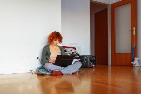 portrait red-haired teen girl sitting on floor in an empty unfurnished room, using tablet, unassembled suitcases standing nearby, concept moving, renting housing for students, studying online at home
