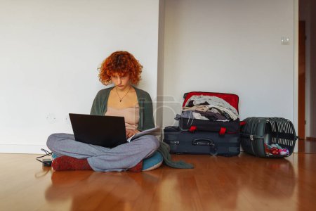 portrait red-haired teen girl sitting on floor in an empty unfurnished room, using tablet, unassembled suitcases standing nearby, concept moving, renting housing for students, studying online at home