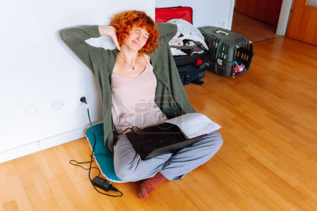 retrato de pelo rojo adolescente chica sentada en el suelo en una habitación vacía sin amueblar, utilizando tableta, maletas sin montar de pie cerca, concepto de movimiento, alquiler de viviendas para los estudiantes, estudiar en línea en casa