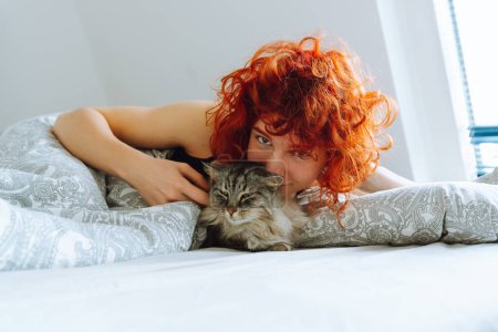 funny red-haired teenage girl, in morning in bed, massages domestic cat, hugs and kisses purring Maine Coon cat. Friendship between pet and its owner