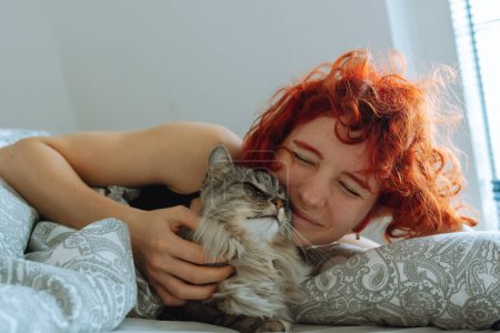 funny red-haired teenage girl, in morning in bed, massages domestic cat, hugs and kisses purring Maine Coon cat. Friendship between pet and its owner