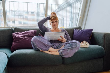 Photo for Portrait middle aged woman, with long red hair tied in bun, sitting on comfortable sofa, wearing loose clothing, using notepad and pen, study at home concept for adults - Royalty Free Image