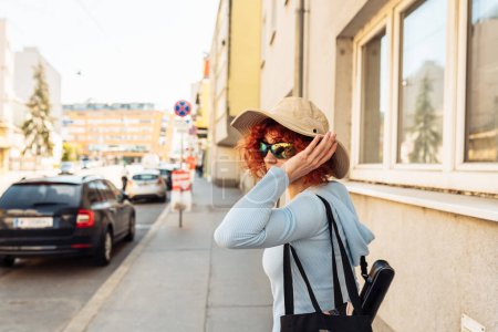 Photo for Portrait young woman, an attractive teenage girl, student, with red curly hair, wearing summer hat and sunglasses, walking along European streets, tourism concept, studenthood, - Royalty Free Image